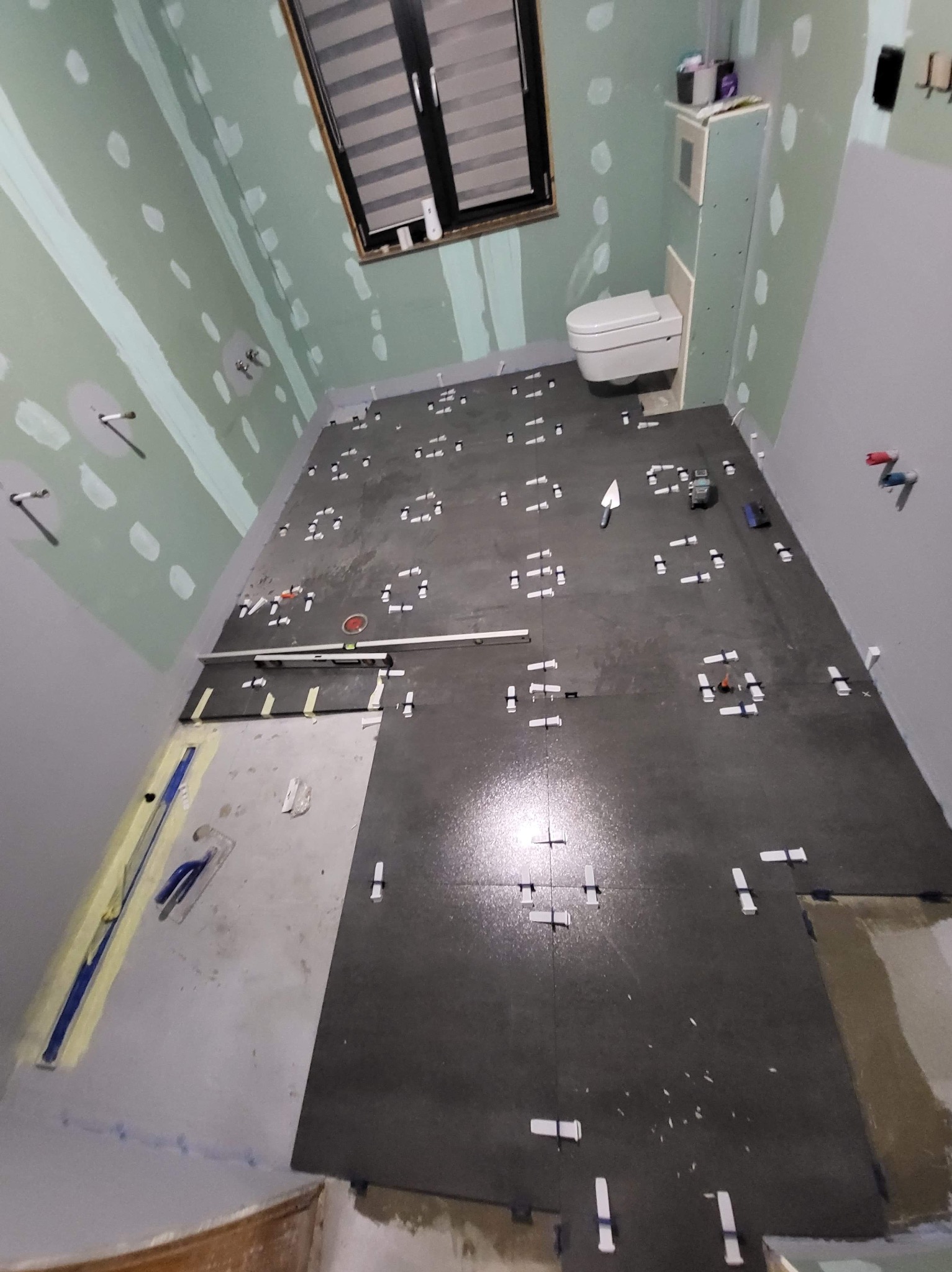 Układanie ciemnych płytek podłogowych w łazience, z białymi krzyżykami dystansowymi. Częściowo przygotowane ściany z płyt karton-gips. Widoczne narzędzia glazurnicze.