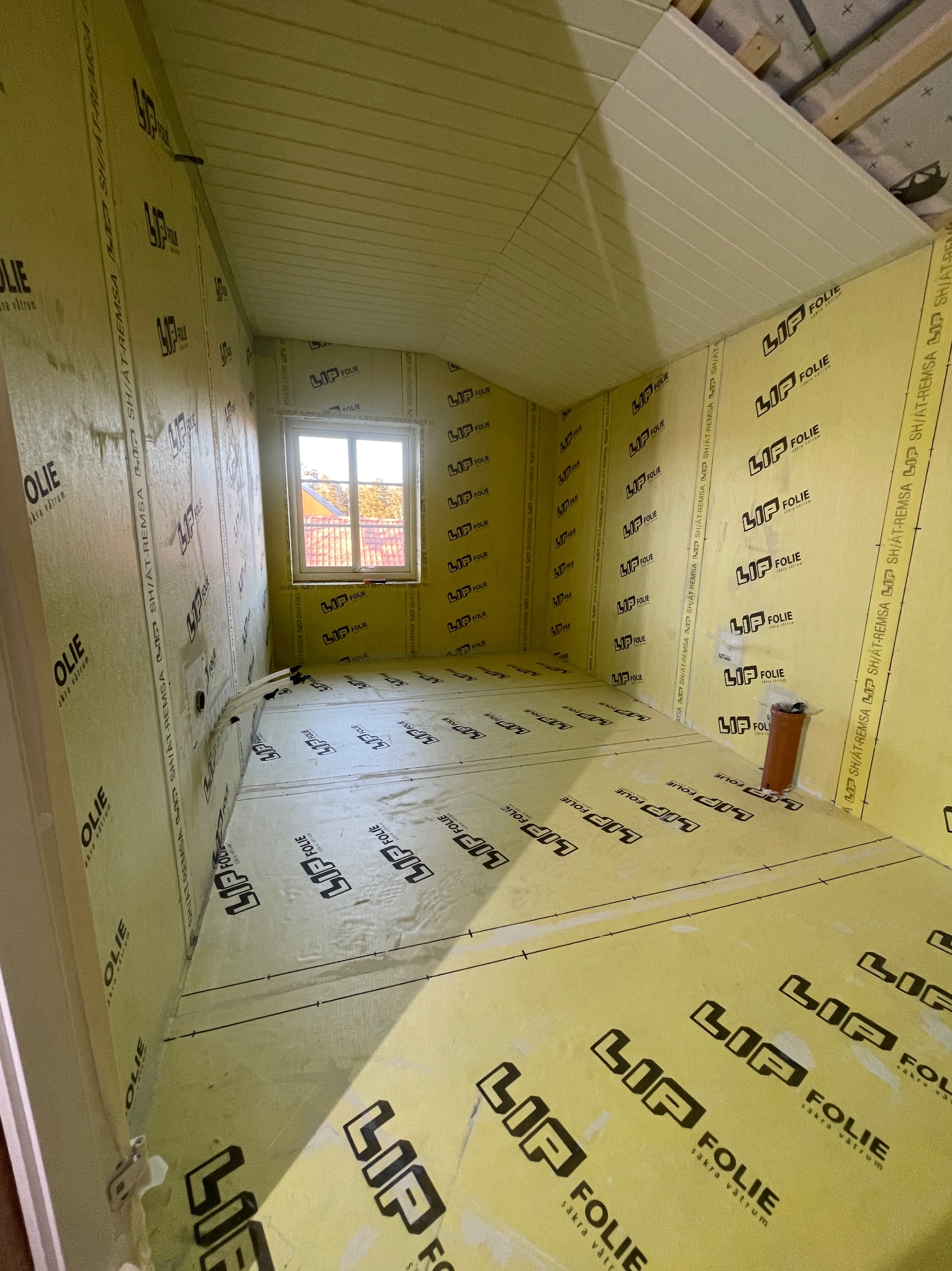 Wnętrze w trakcie prac wykończeniowych: ściany i podłoga pokryte żółtą folią izolacyjną z czarnym nadrukiem, widoczne okno i fragment sufitu.