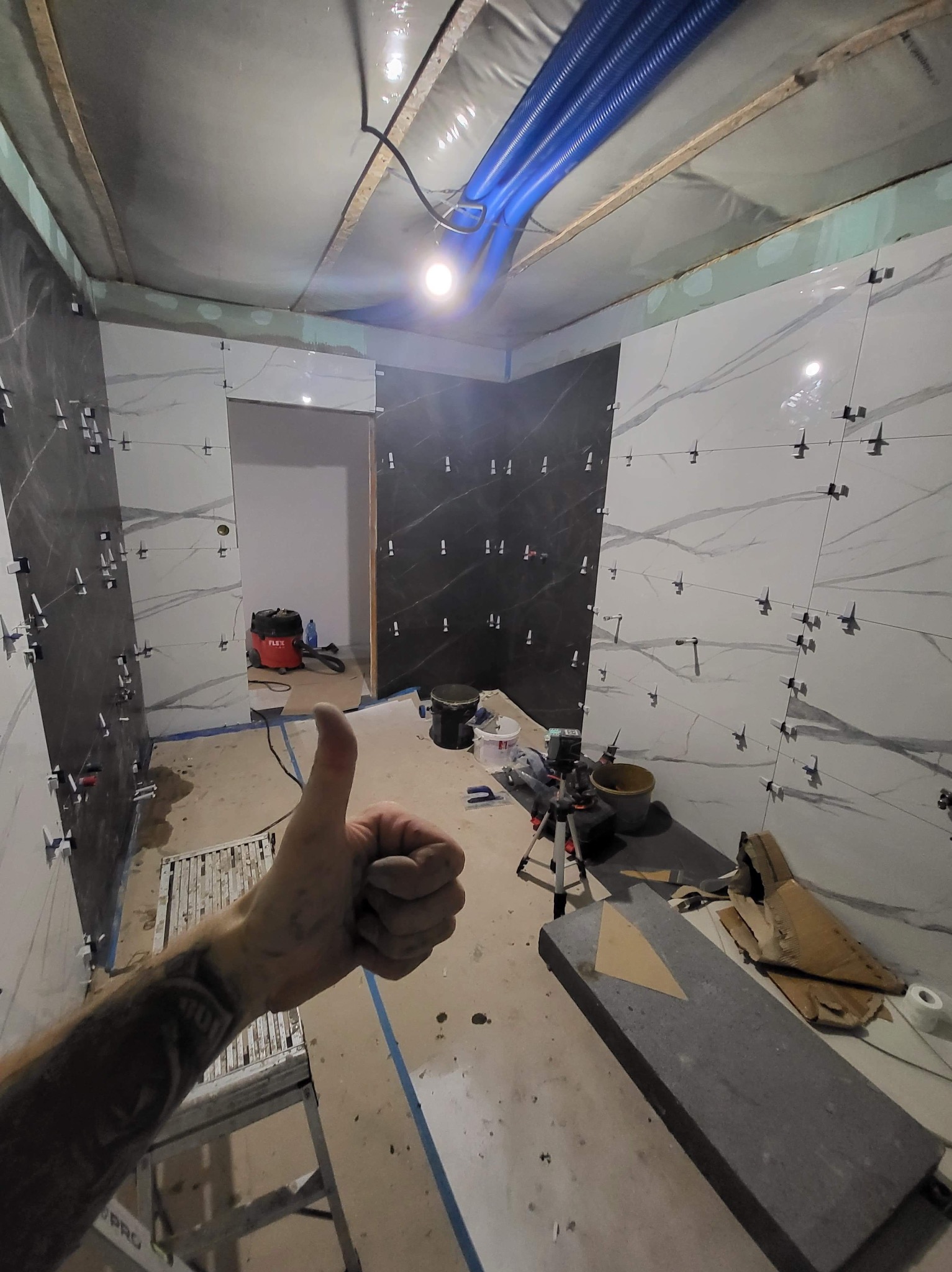 Układanie płytek w łazience z użyciem krzyżyków dystansowych. Widoczna ręka z kciukiem w górę. W tle narzędzia glazurnicze i częściowo ułożone płytki imitujące marmur.