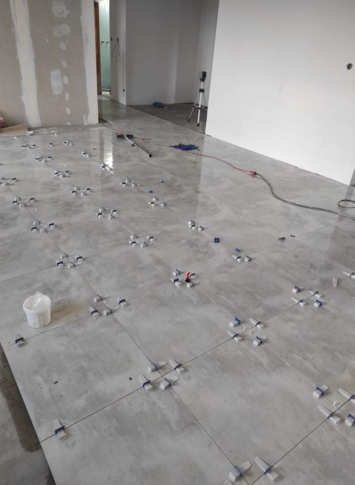 Układanie szarej, matowej terakoty w salonie z użyciem krzyżyków dystansowych i poziomicy. Widoczny biały pojemnik i przewody na podłodze.