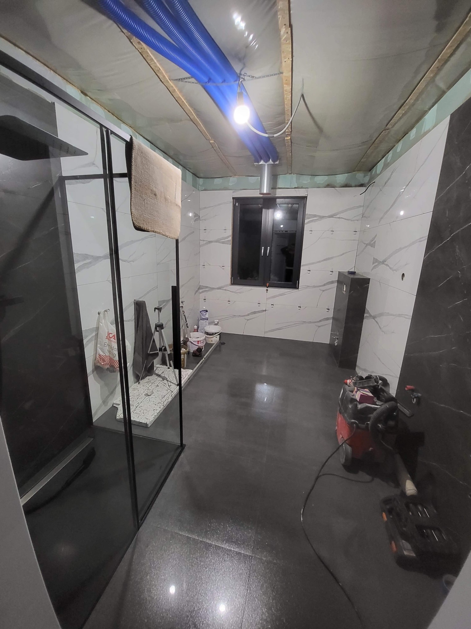 Wnętrze łazienki w trakcie wykończenia: widoczne płytki imitujące marmur, kabina prysznicowa z czarnymi profilami, narzędzia i niebieskie rury wentylacyjne pod sufitem.