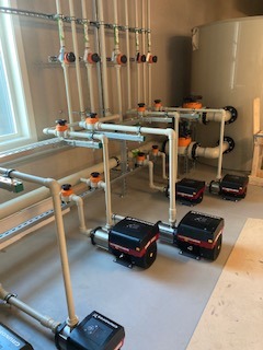 Kompleksowa instalacja hydrauliczna: szereg pomp obiegowych Grundfos z rurami, zbiornik wyrównawczy i liczne zawory, zamontowane schludnie na ścianie w jasnym pomieszczeniu.