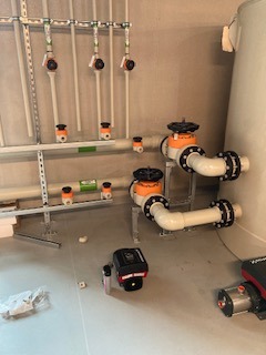 Białe rury z zaworami i pompami zamontowane na ścianie i podłodze w pomieszczeniu technicznym. Widoczne elementy instalacji wodnej i grzewczej.