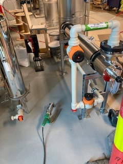 Instalacja przemysłowa z rur PVC i elementami ze stali nierdzewnej, widoczne złącza i zawory, na posadzce leżą narzędzia. Ujęcie z bliska, oświetlenie przemysłowe.