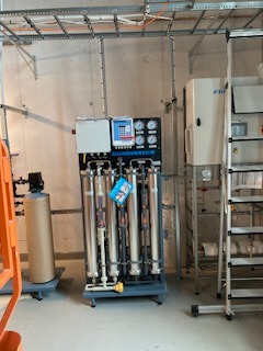 Filtracja wody: stalowa instalacja z manometrami i cylindrami filtracyjnymi w pomieszczeniu technicznym. Widoczne rury, zawory i podłączenia elektryczne.