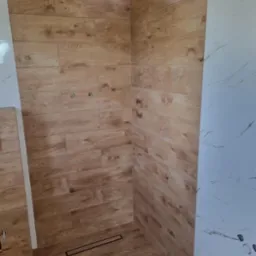 Kabina prysznicowa wykończona płytkami imitującymi drewno i marmur, z widocznym odpływem liniowym. Świeżo położone płytki, widoczne otwory na armaturę.