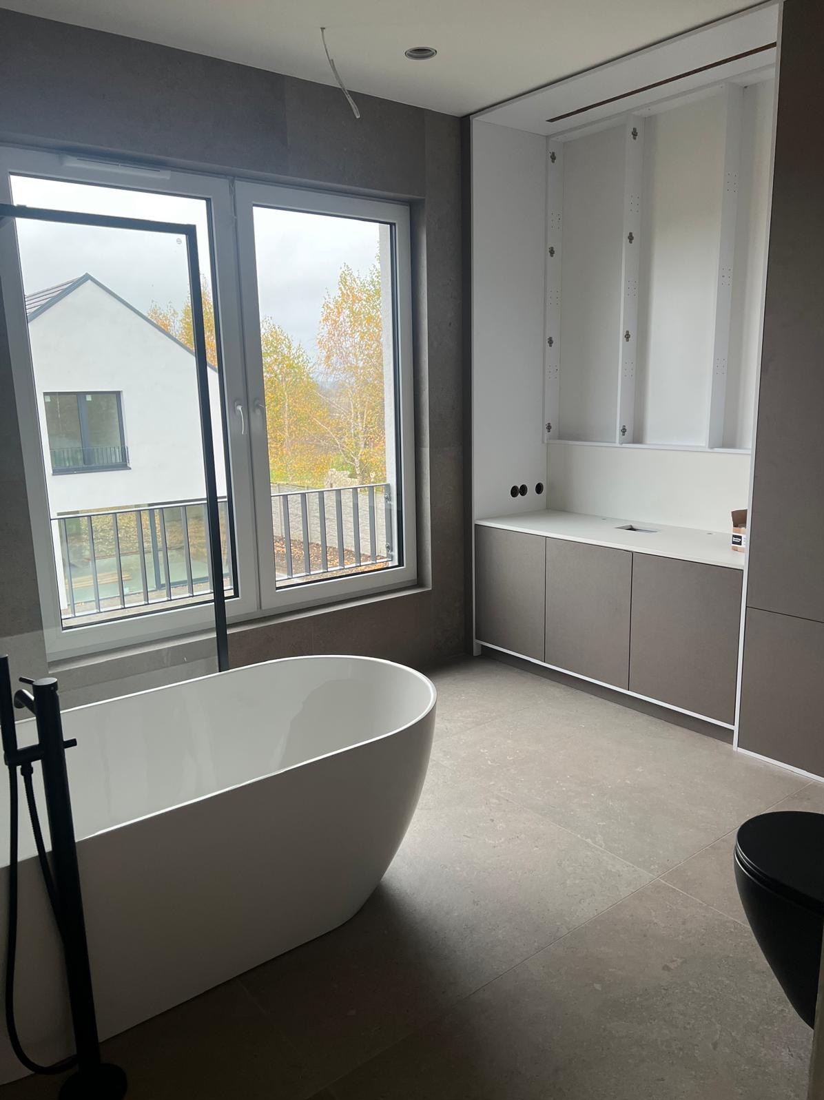 Minimalistyczna łazienka w szarościach z wolnostojącą wanną i widokiem na jesienne drzewa za oknem. Uwagę zwraca nowoczesna zabudowa meblowa.