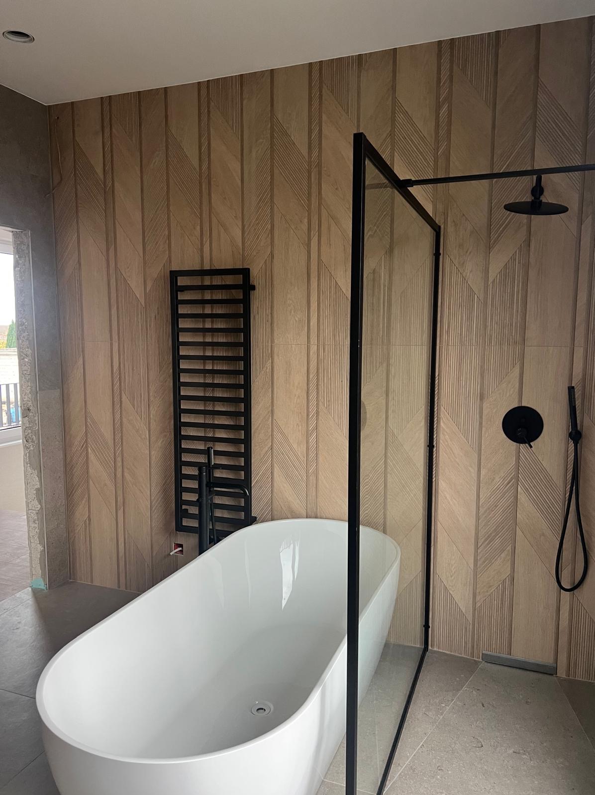 Nowoczesna łazienka z wolnostojącą wanną, czarną armaturą i grzejnikiem na tle ściany wyłożonej płytkami imitującymi drewno w geometryczny wzór.