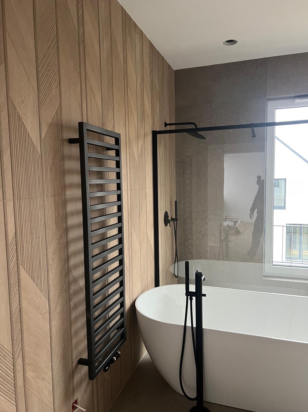 Nowoczesna łazienka z wolnostojącą wanną, czarną armaturą i grzejnikiem drabinkowym na ścianie wyłożonej płytkami imitującymi drewno w geometryczny wzór.