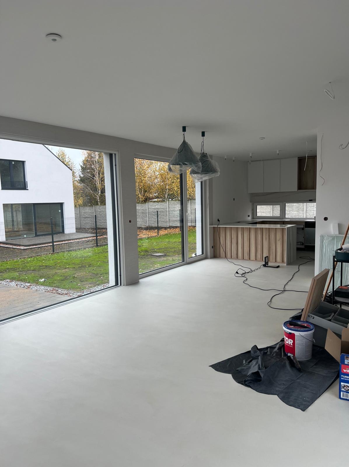 Jasne wnętrze domu z dużymi oknami po wylaniu posadzki, widoczne narzędzia i materiały budowlane, surowy stan wykończenia. Minimalistyczny styl.