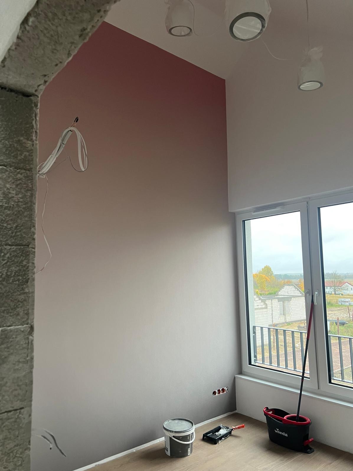 Ściana pomalowana gradientem różu, widoczne narzędzia malarskie, niezabezpieczone kable i surowy kamienny łuk. Widok z okna na budowę.