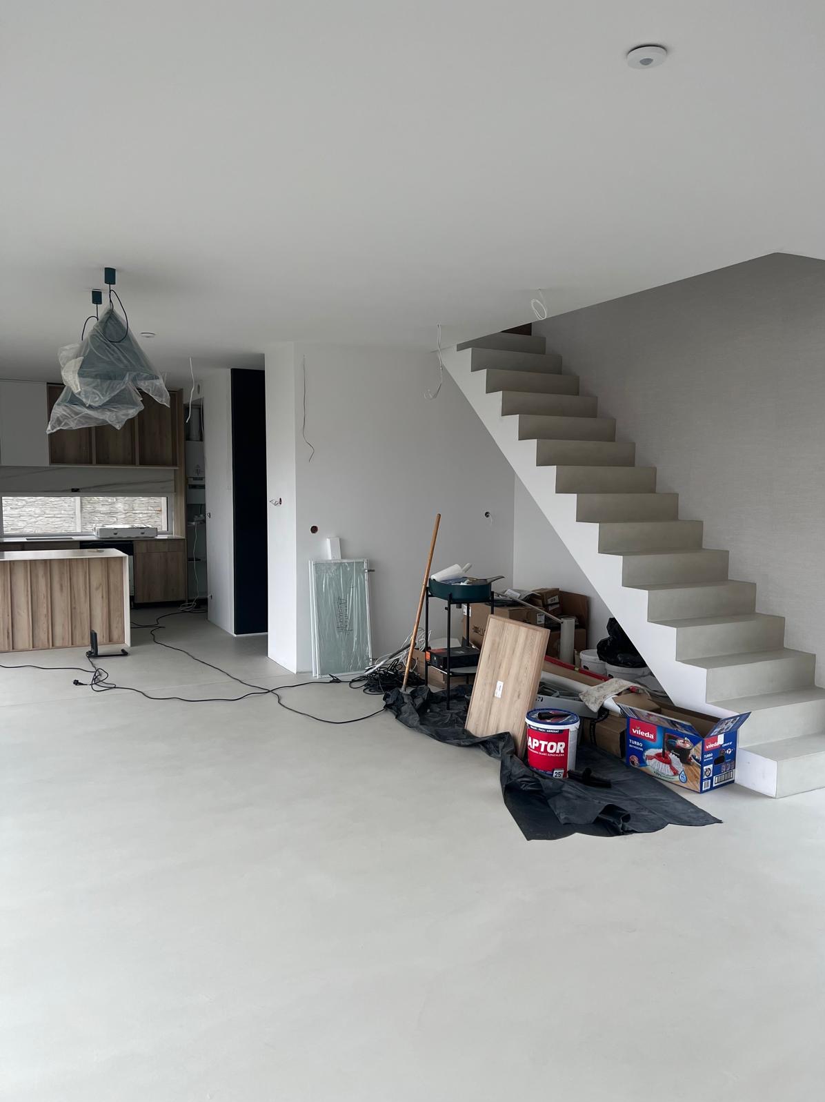 Wnętrze w trakcie prac wykończeniowych: białe ściany, surowe schody, widoczne narzędzia i materiały budowlane, w tle zabudowa kuchenna pod folią ochronną.