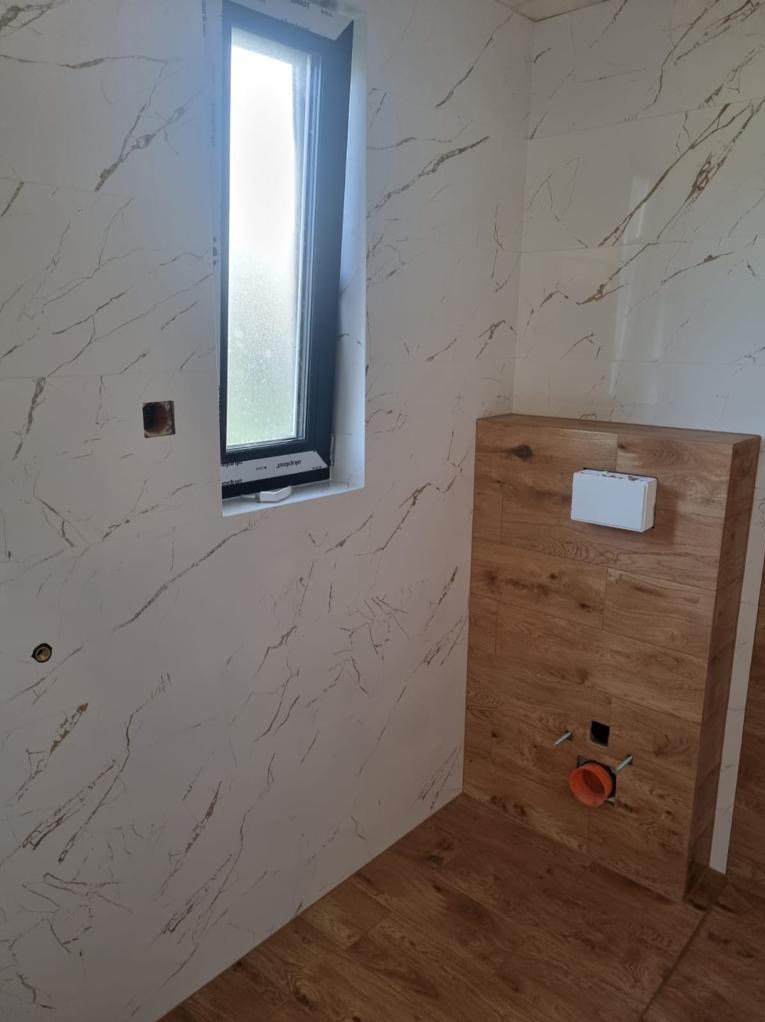 Wykończona łazienka z marmurowymi płytkami ściennymi, drewnopodobną obudową stelaża WC i oknem. Widoczne instalacje hydrauliczne.
