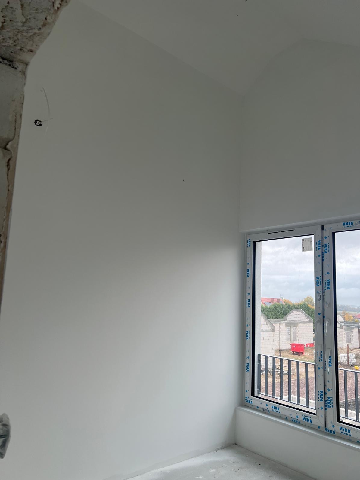 Gładka, biała ściana w rogu pokoju z oknem Veka. Widok na budowę za oknem. Fragment surowej ściany po lewej stronie. Minimalistyczne wnętrze w trakcie wykończenia.