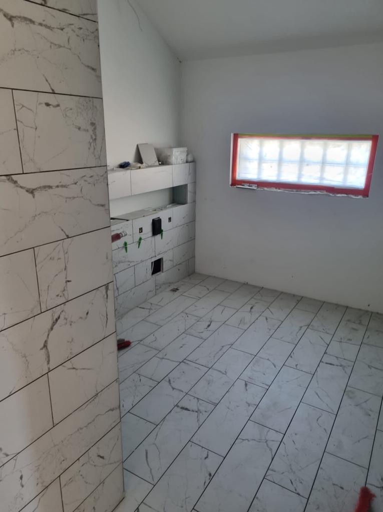 Wnętrze łazienki w trakcie remontu: białe płytki imitujące marmur na ścianach i podłodze, widoczne otwory na instalacje, okno z czerwoną ramą zabezpieczone folią.