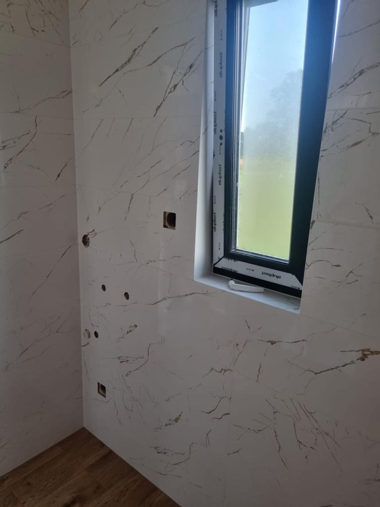 Ściana wyłożona białymi płytkami imitującymi marmur z widocznymi otworami instalacyjnymi i oknem z czarną ramą, fragment drewnianej podłogi w rogu.