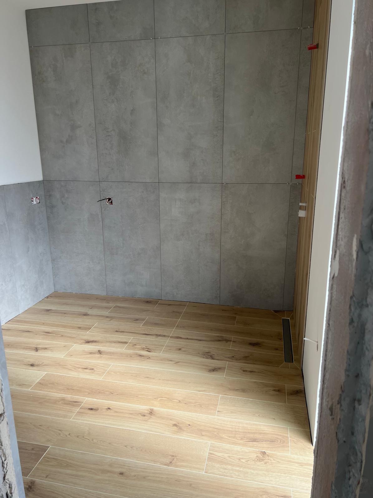 Wykończona łazienka z szarymi płytkami ściennymi i drewnopodobną podłogą. Widoczne detale jak listwa odpływowa i system poziomowania płytek.