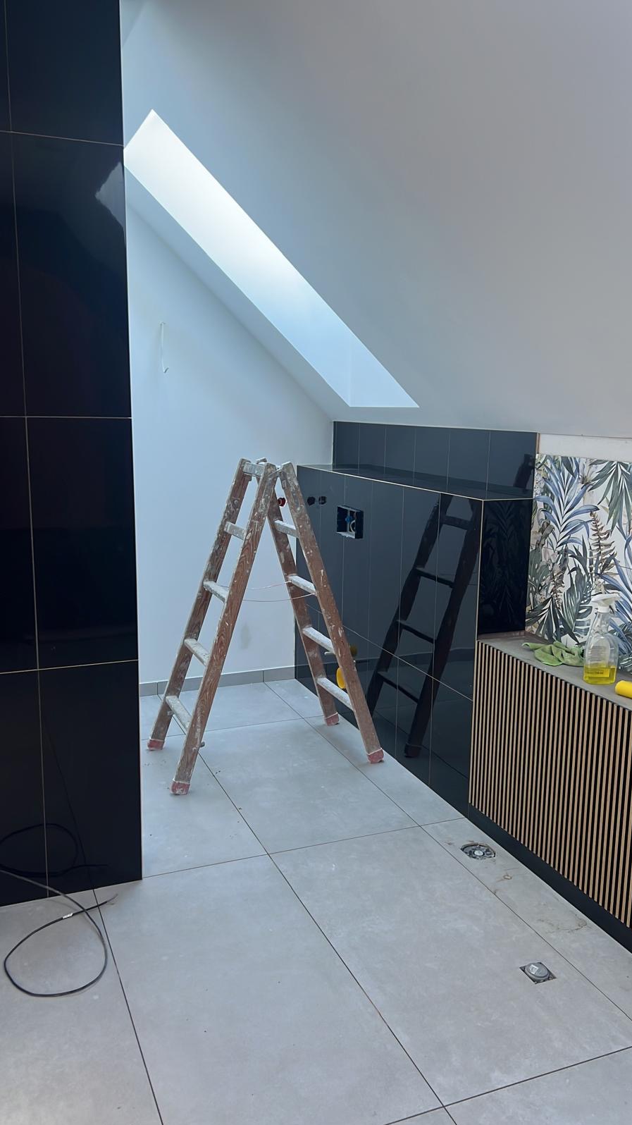 Remont łazienki na poddaszu: czarne płytki ścienne, drabina malarska, częściowo ułożone płytki z wzorem roślinnym. Widoczne okno dachowe.