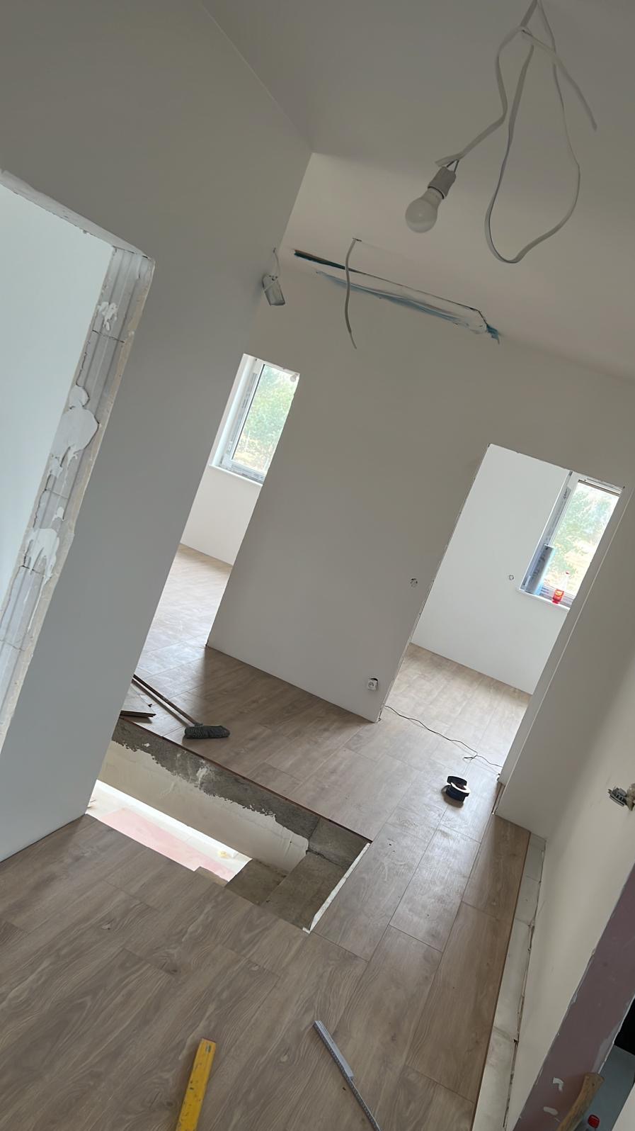 Jasne wnętrze w trakcie remontu: świeżo położona podłoga, surowe ściany, okna w tle. Widoczne narzędzia i niedokończone prace. Dom w trakcie modernizacji.