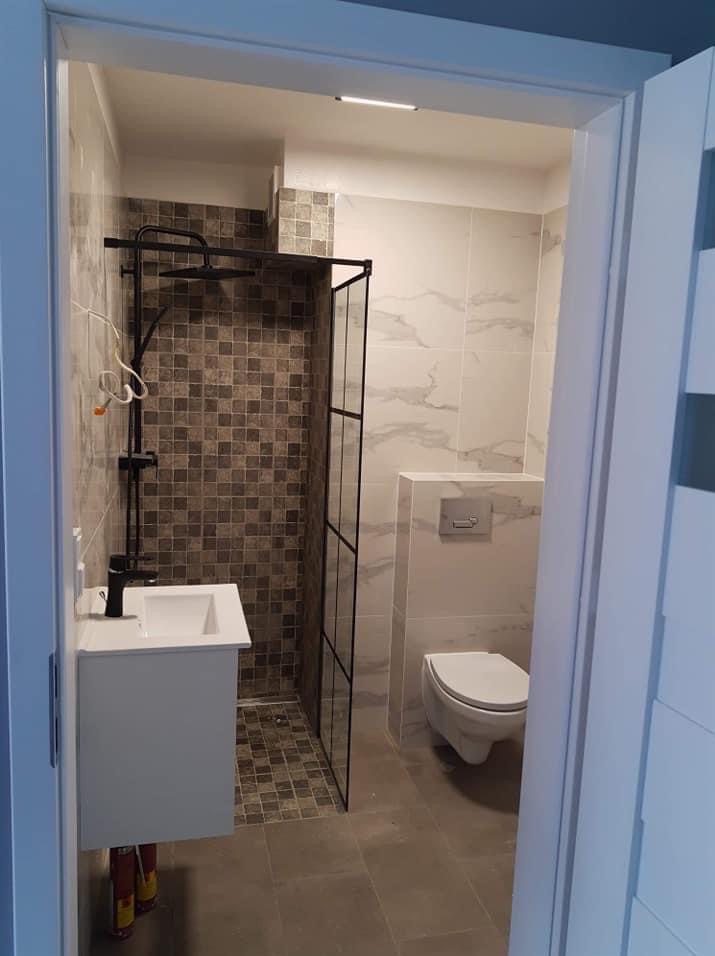 Wykończona łazienka z kabiną prysznicową typu walk-in, umywalką z szafką i toaletą. Dominuje szara kolorystyka płytek i czarne akcenty armatury.