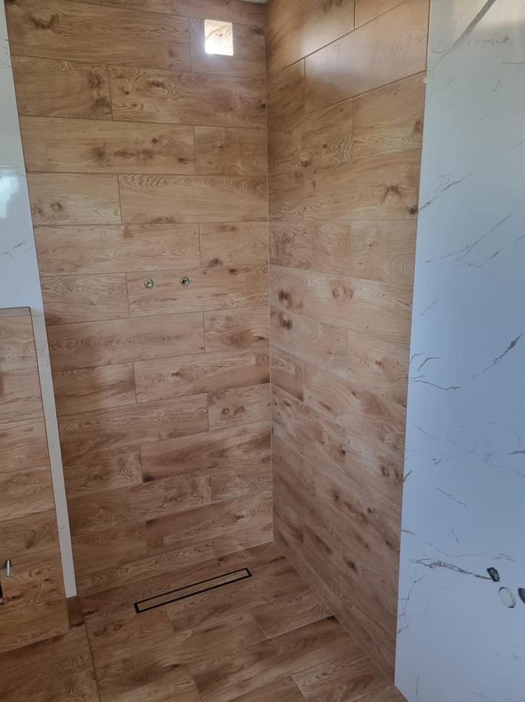 Kabina prysznicowa wykończona płytkami imitującymi drewno i marmur, z widocznym odpływem liniowym. Świeżo położone płytki, widoczne otwory na armaturę.