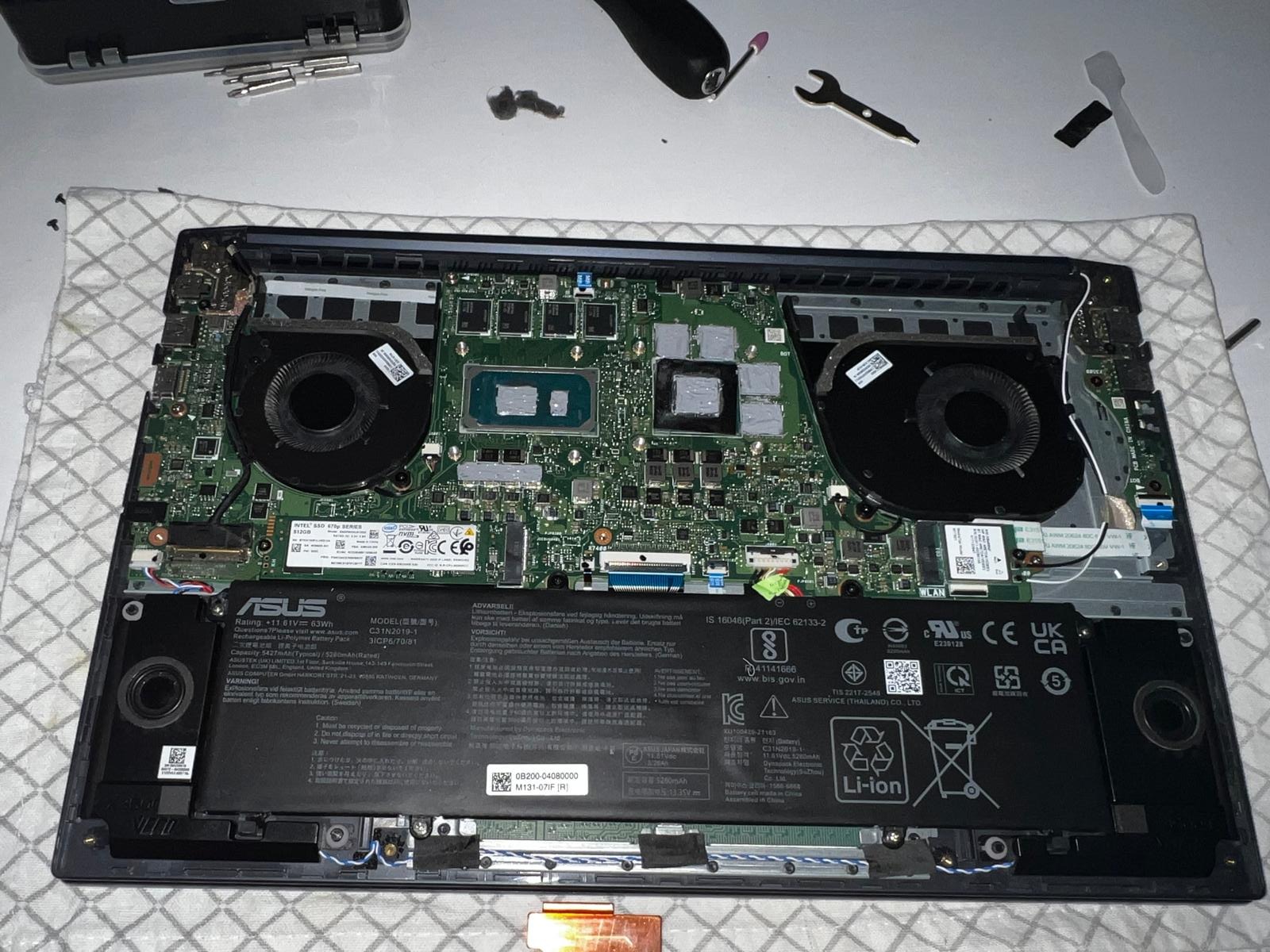 Otwarty laptop ASUS z widoczną płytą główną, układem chłodzenia i baterią. Widoczne narzędzia sugerują naprawę lub konserwację. Detaliczne ujęcie elektroniki.