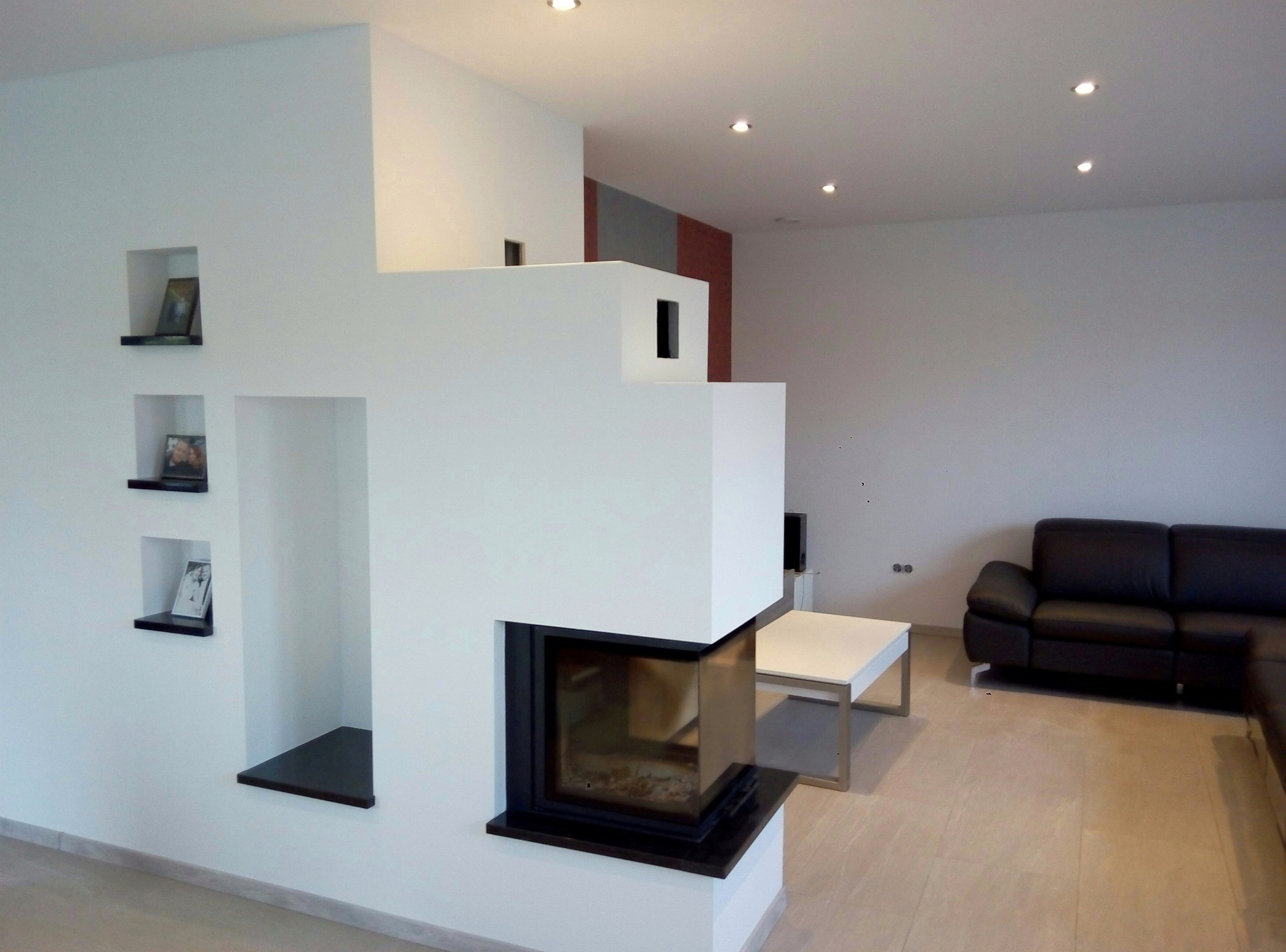 Minimalistyczny salon z białymi ścianami, wbudowanym kominkiem i półkami wnękowymi. Nowoczesne wykończenie wnętrza, jasna podłoga, ciemna sofa.