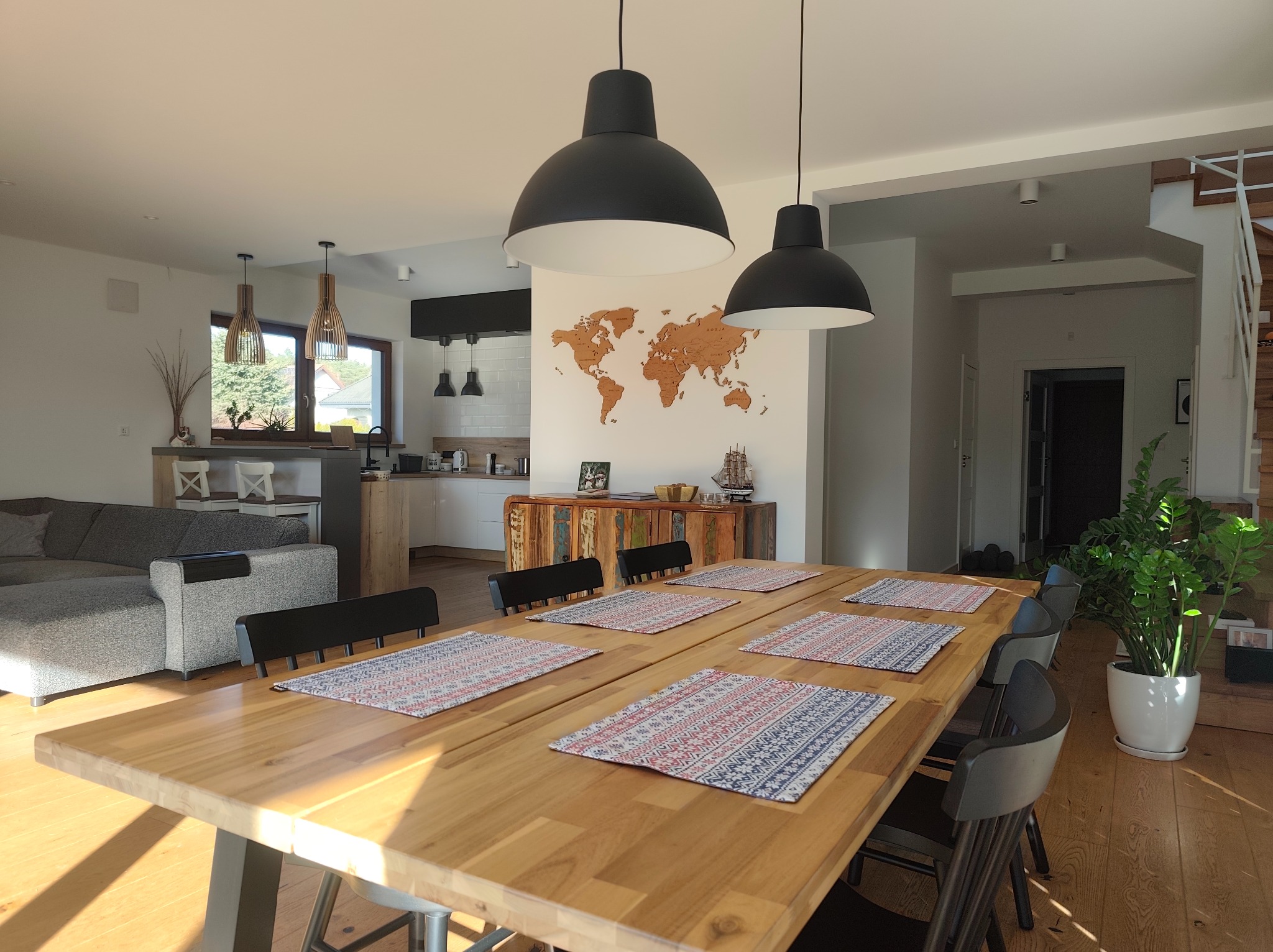 Jasne, przestronne wnętrze domu z drewnianym stołem jadalnym, czarnymi krzesłami i dekoracyjną mapą świata na ścianie. Nowoczesne lampy wiszące i parkiet dodają elegancji.
