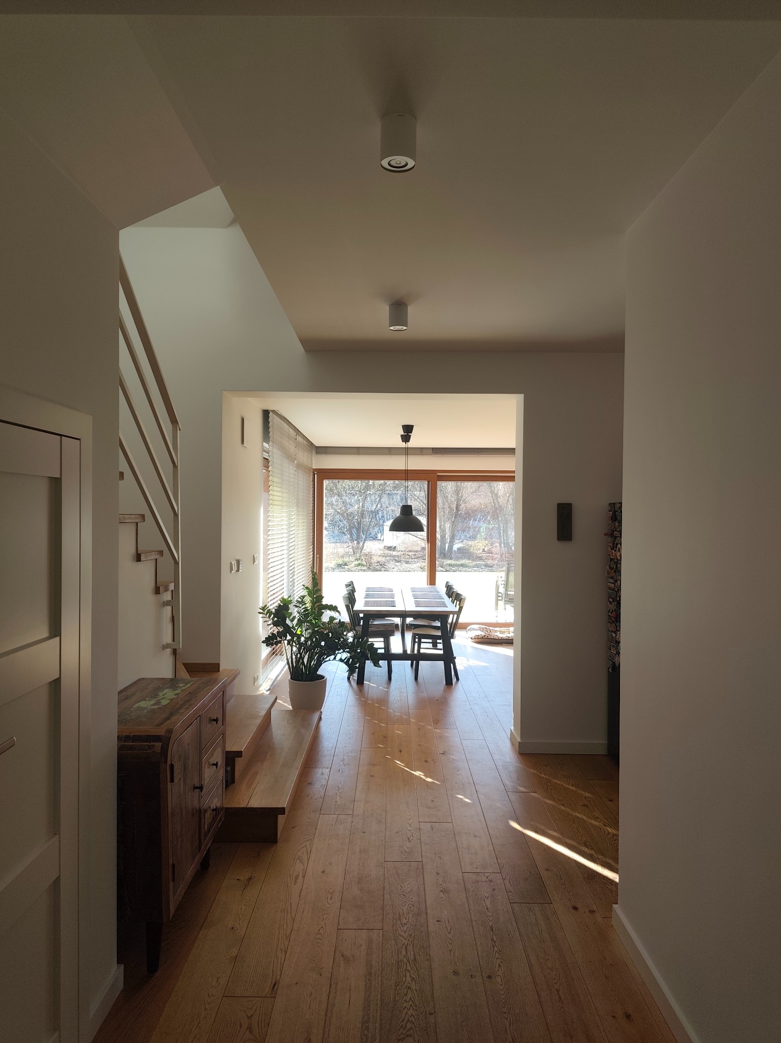 Jasne wnętrze domu z drewnianą podłogą i schodami. Widok na jadalnię z dużym oknem. Minimalistyczny design, neutralna kolorystyka ścian, roślina doniczkowa.