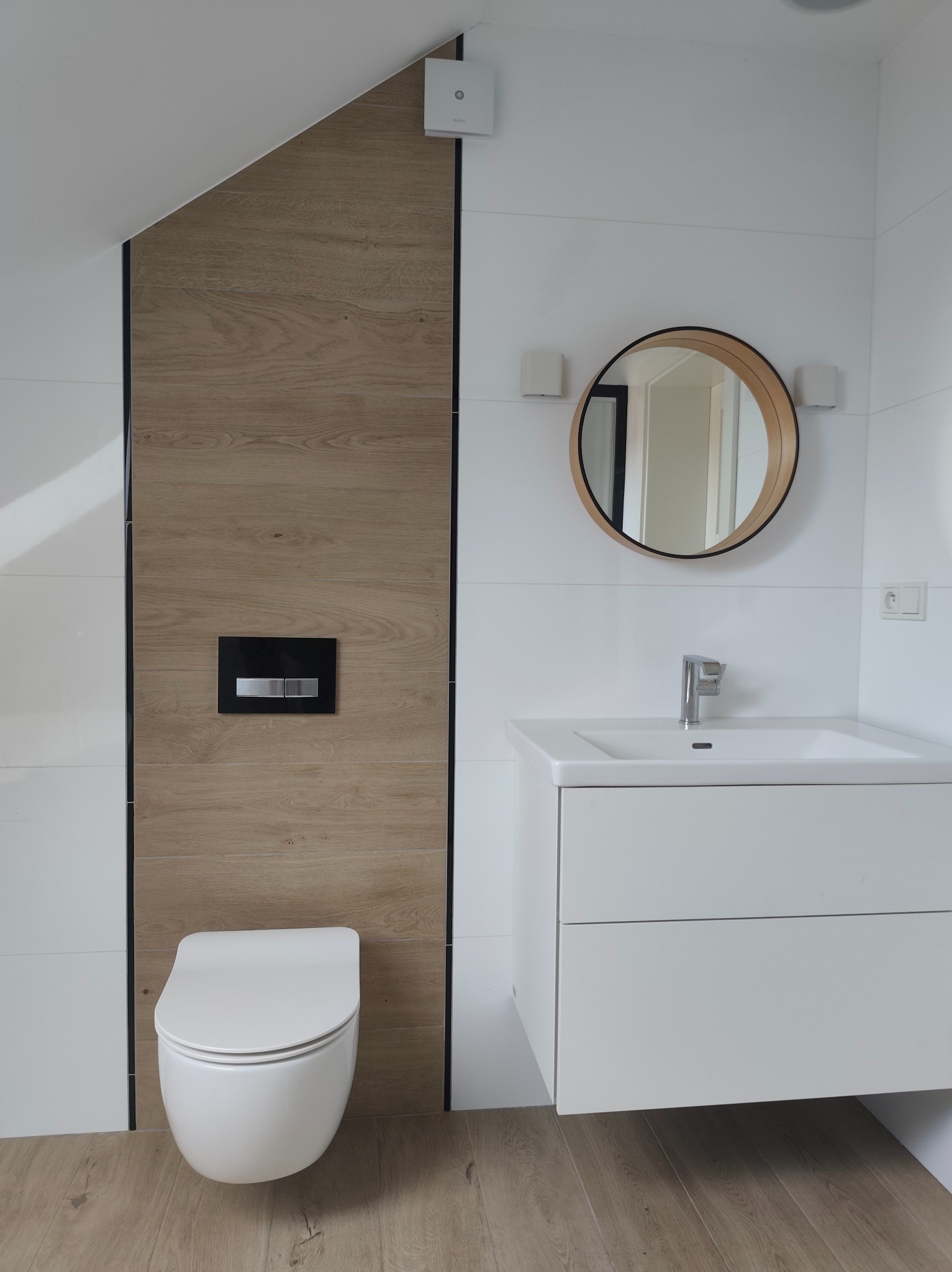 Nowoczesna łazienka na poddaszu z białymi płytkami i drewnianymi akcentami. Okrągłe lustro nad białą szafką z umywalką. Podwieszana toaleta na tle drewnopodobnych płytek.