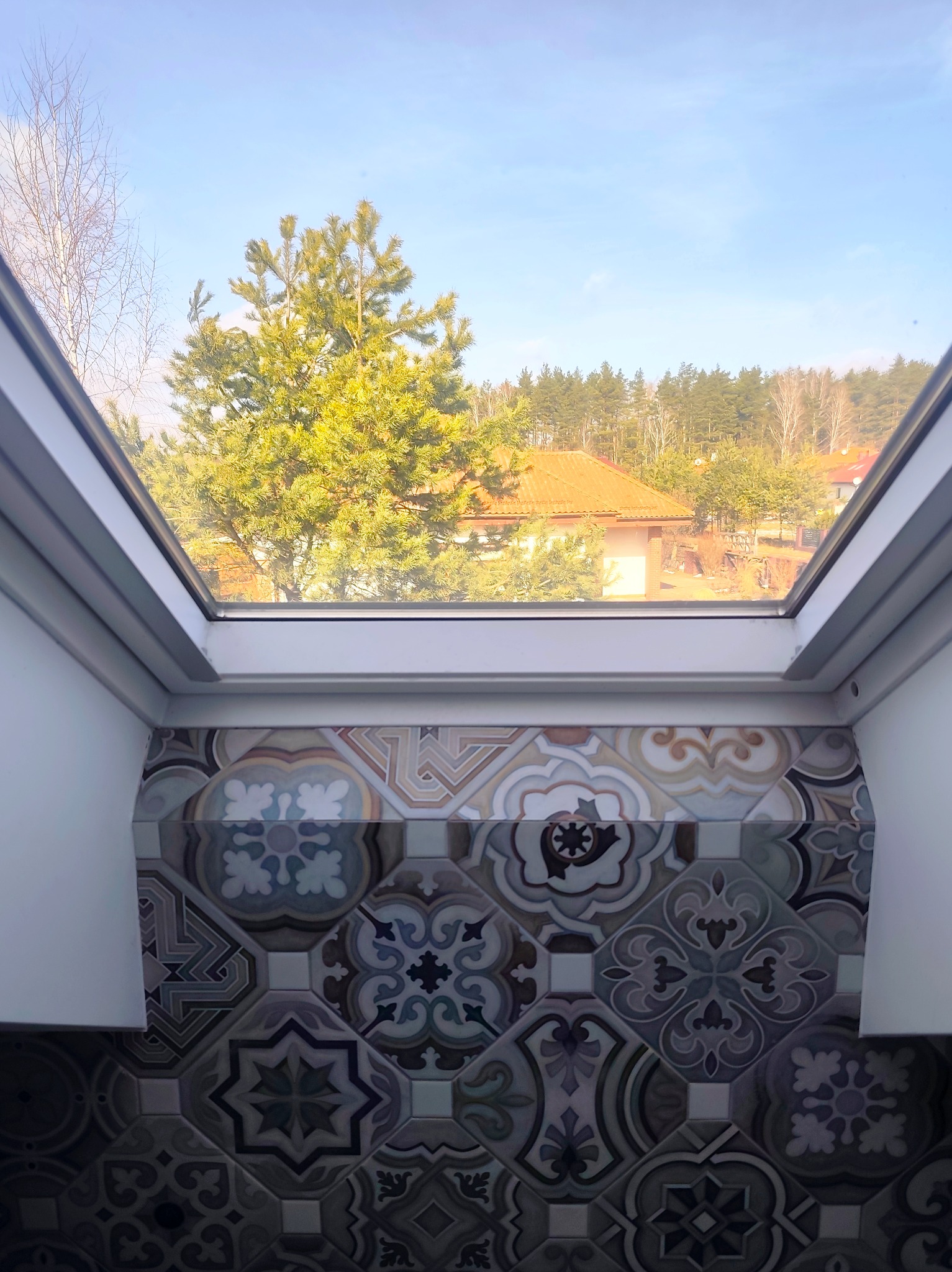 Wnętrze z oknem dachowym, ukazujące dekoracyjne płytki ścienne w odcieniach szarości i beżu. Za oknem widok na drzewa i dom z pomarańczowym dachem.
