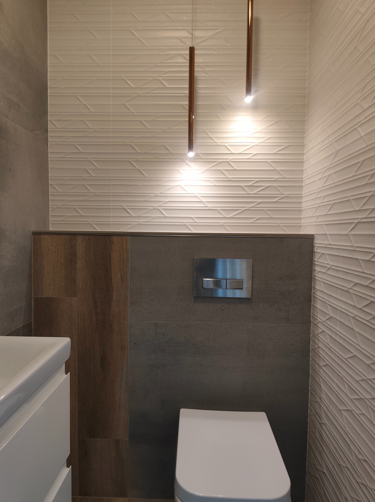 Nowoczesna toaleta z podwieszaną miską WC, szarą i drewnopodobną okładziną oraz dekoracyjnymi, białymi płytkami 3D i minimalistycznym oświetleniem.