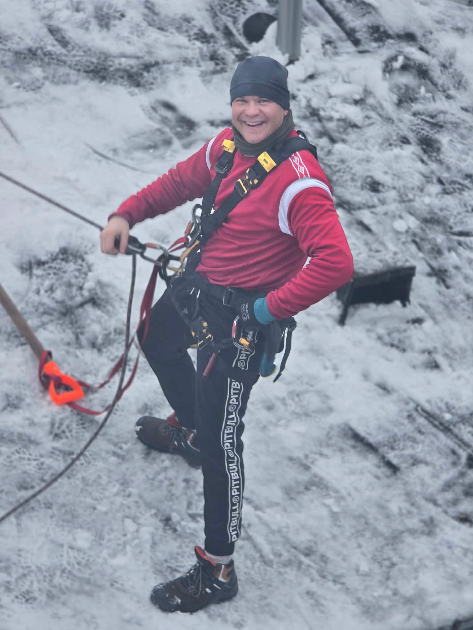 Uśmiechnięty mężczyzna w uprzęży, na pochyłym, ośnieżonym dachu. Widoczne liny i narzędzia. Praca na wysokości w zimowych warunkach.