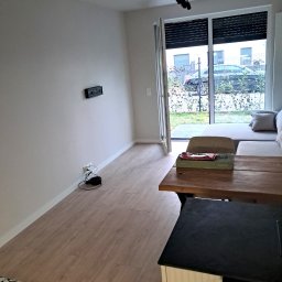 KAVER SPÓŁKA Z OGRANICZONĄ ODPOWIEDZIALNOŚCIĄ - Jasne wnętrze z drewnianą podłogą i częściowo odsłoniętymi roletami. Widoczny fragment sofy i stolika. Na ścianie uchwyt pod telewizor, brak sprzętu.