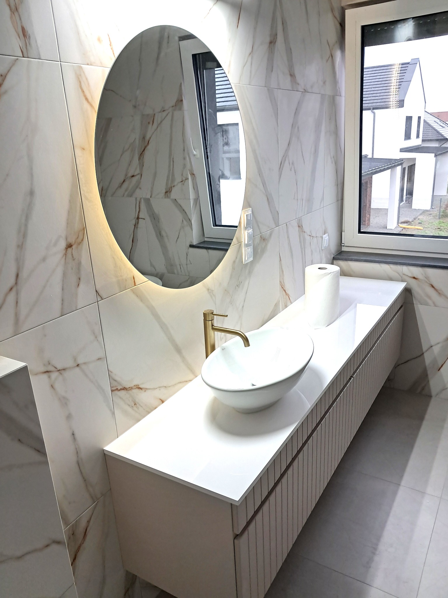 Wykończona łazienka w Katowicach: marmurowe płytki, owalne lustro z podświetleniem LED i biała umywalka nablatowa z baterią w kolorze złota.