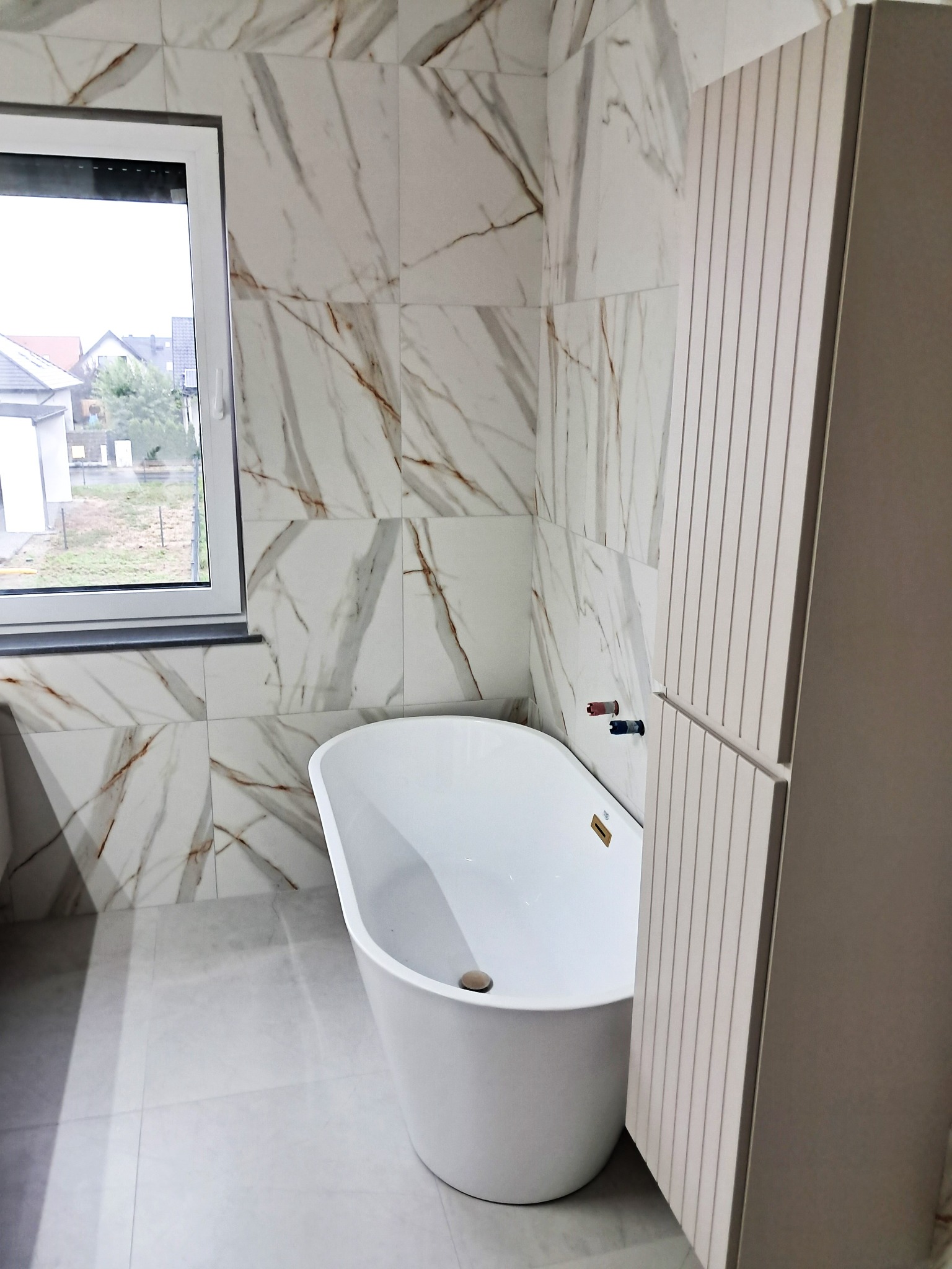 Nowoczesna łazienka: biała wanna wolnostojąca pod oknem, marmurowe płytki ścienne z brązowymi żyłami, szara podłoga i zabudowa meblowa z pionowymi frezami.