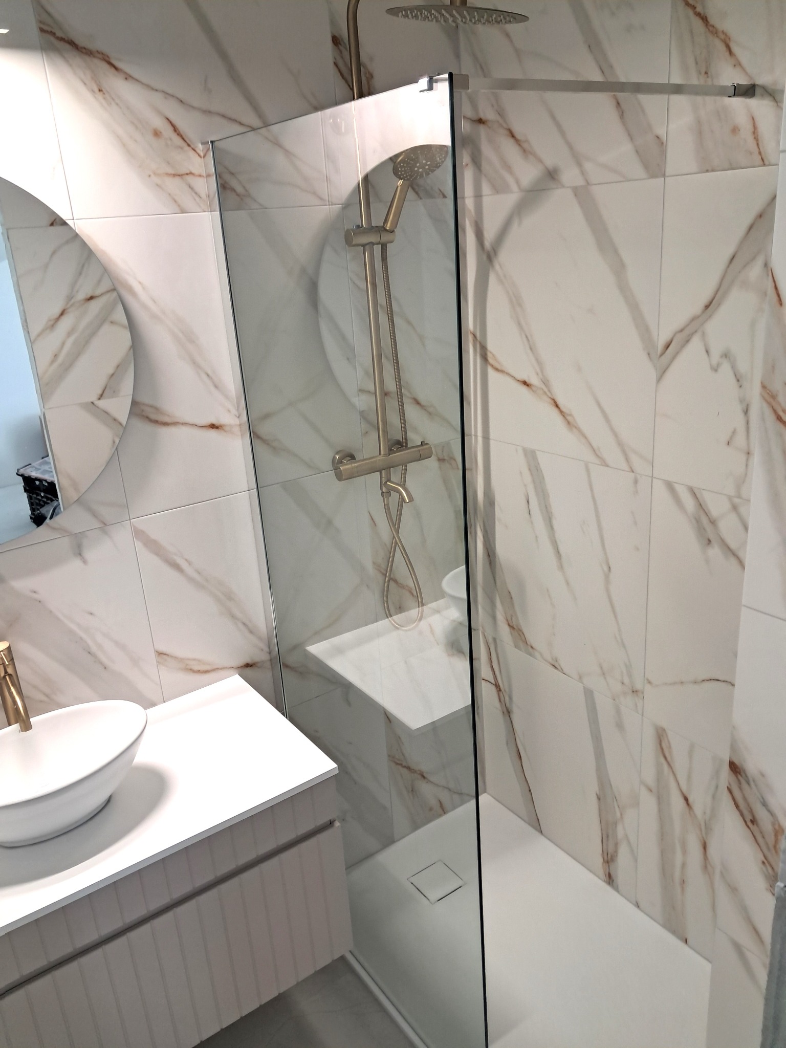 Nowoczesna łazienka z kabiną prysznicową typu walk-in, umywalką nablatową i lustrem w okrągłej ramie. Płytki imitujące marmur z brązowymi żyłami.