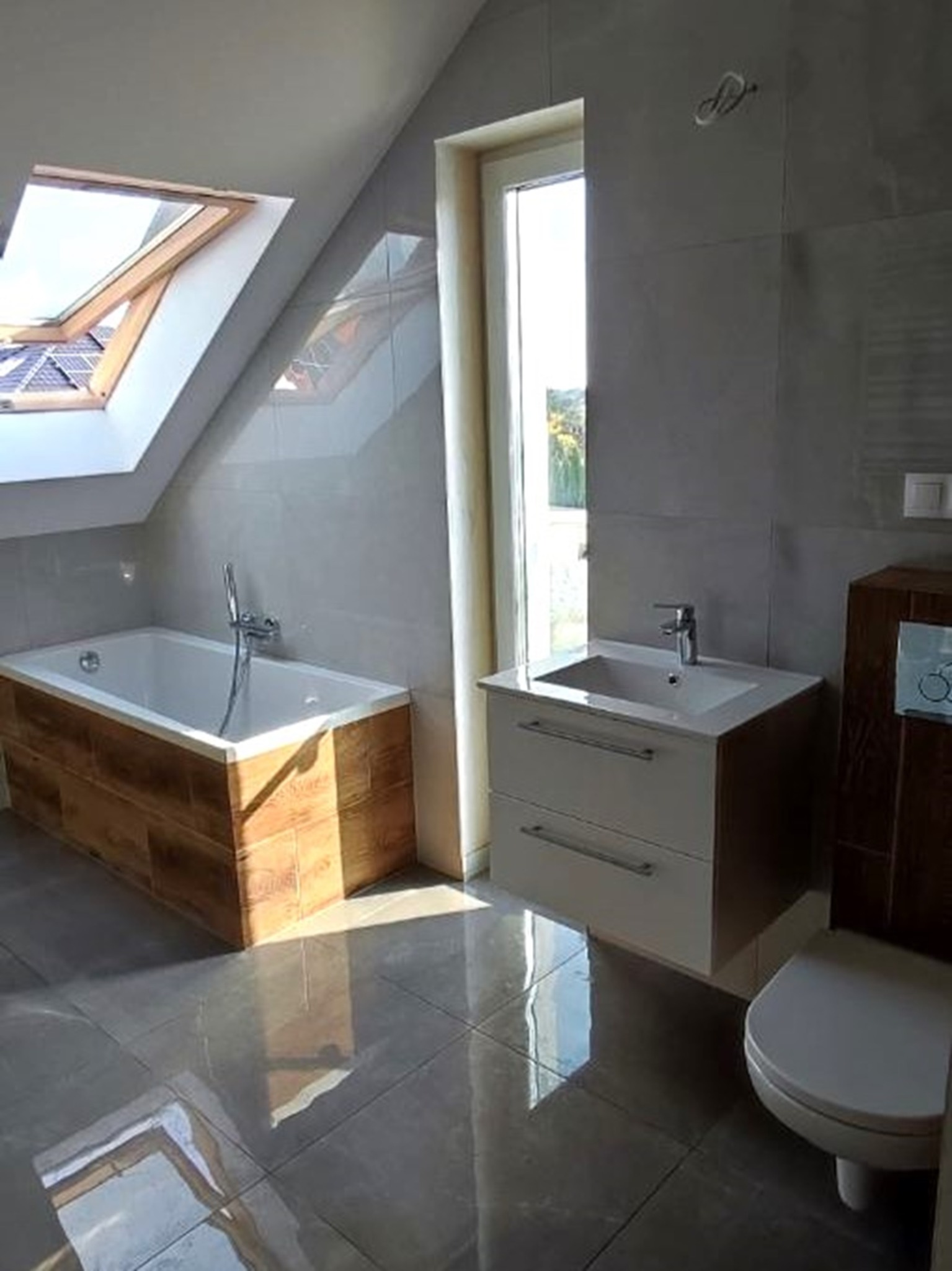 Nowoczesna łazienka na poddaszu z oknem dachowym: wanna obudowana drewnem, umywalka z szafką i toaleta. Szare płytki na ścianach i podłodze. Jasne, naturalne światło.