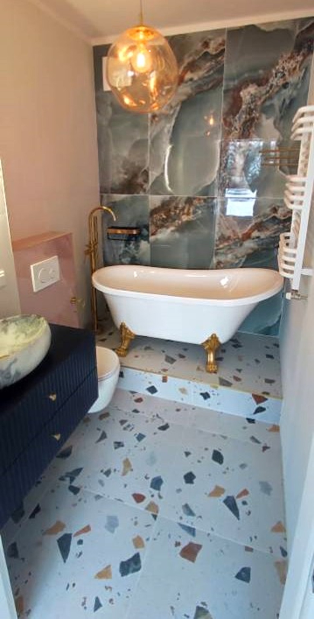 Elegancka łazienka z wolnostojącą wanną na złotych nóżkach, płytkami imitującymi onyks i podłogą lastryko. Złota armatura i dekoracyjna lampa.