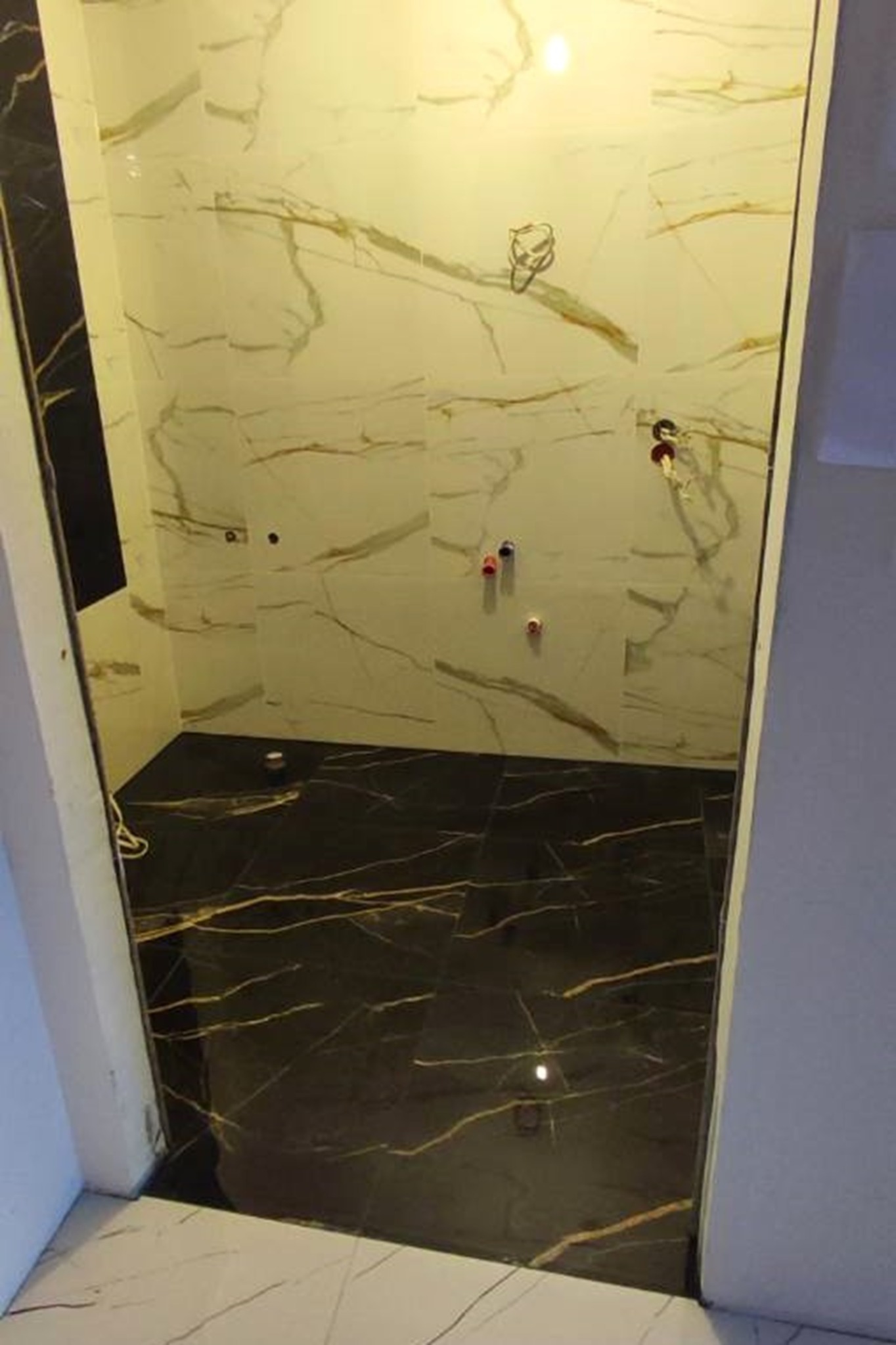 Wykończona kabina prysznicowa z czarno-białymi marmurkowymi płytkami, widoczne przyłącza wodne. Ciemna podłoga kontrastuje z jasnymi ścianami.