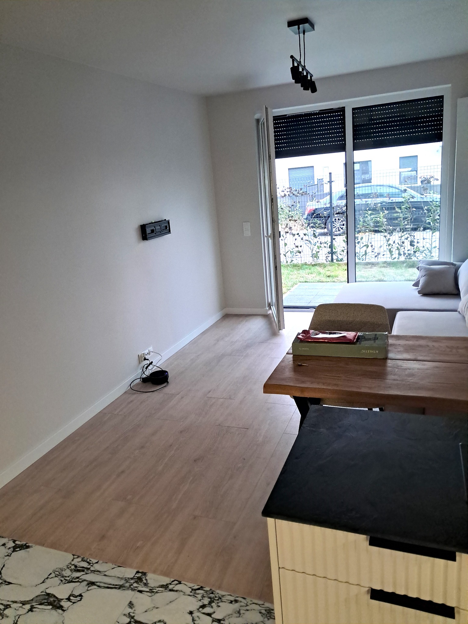 Jasne wnętrze z drewnianą podłogą i częściowo odsłoniętymi roletami. Widoczny fragment sofy i stolika. Na ścianie uchwyt pod telewizor, brak sprzętu.