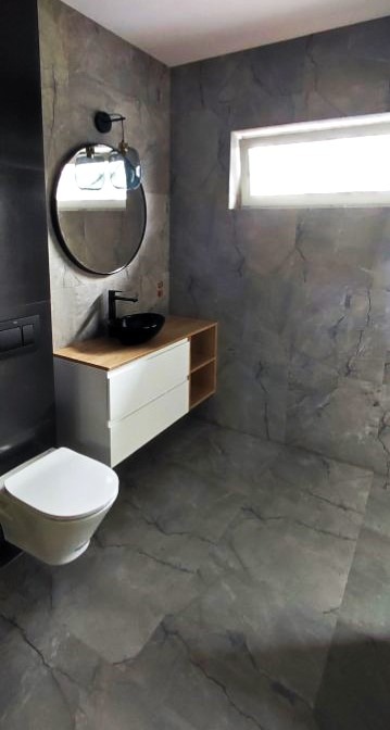 Nowoczesna łazienka w szarościach z okrągłym lustrem, umywalką nablatową i podwieszaną toaletą. Ściany i podłoga wykończone płytkami imitującymi marmur.