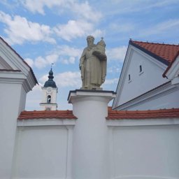 WE-NA - Kamienna figura świętego na postumencie, biały mur z czerwoną dachówką i wieża kościelna w tle pod błękitnym niebem.