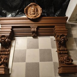 WE-NA - Dekoracyjny, drewniany portal kominkowy z rzeźbioną płaskorzeźbą twarzy, ustawiony na podłodze z płytek w odcieniach szarości, pod ścianą w trakcie prac remontowych.