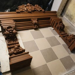 WE-NA - Drewniany, rzeźbiony portal kominkowy w odcieniach brązu z dekoracyjnymi elementami, leżący na podłodze wyłożonej kwadratowymi płytkami.