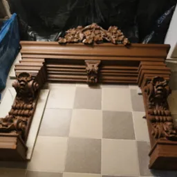 Odrestaurowany, brązowy portal kominkowy z rzeźbionymi elementami roślinnymi i motywami twarzy, ustawiony na podłodze z kwadratowych płytek.