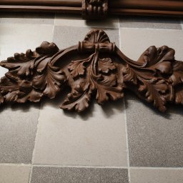 WE-NA - Detal dekoracyjny z brązowego drewna, prawdopodobnie fragment kominka lub pieca kaflowego, na tle wzorzystych płytek. Widoczne elementy rzeźbione w kształcie liści.