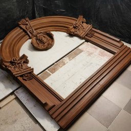 WE-NA - Rozłożone elementy dekoracyjne z drewna, prawdopodobnie fragment kominka lub portalu, z rzeźbionymi detalami i motywem twarzy, na tle podłogi w kratkę.