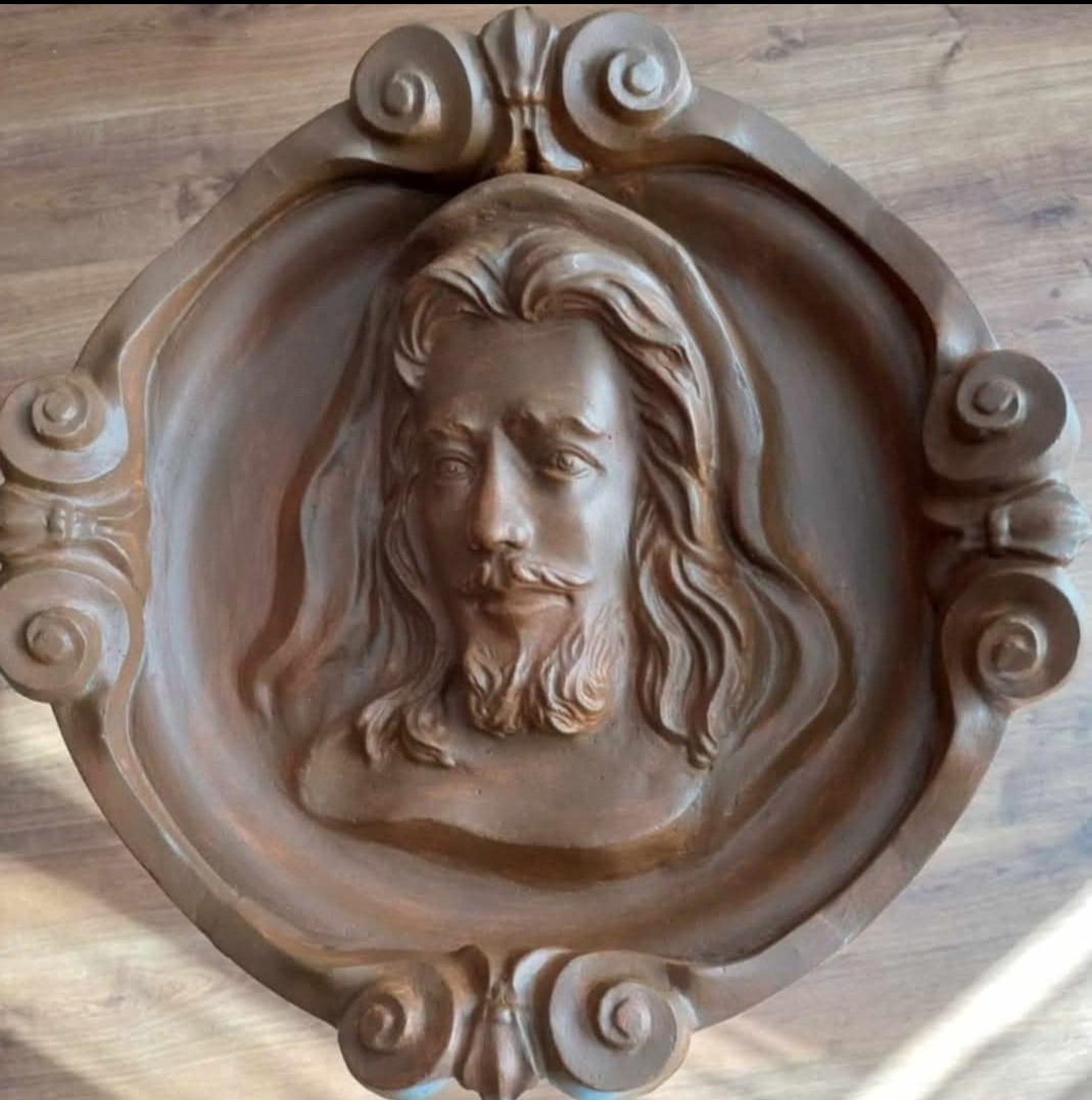 Owalna płaskorzeźba z wizerunkiem mężczyzny z brodą i długimi włosami, w ozdobnej, stylizowanej ramie, wykonana z terakoty lub innego materiału ceramicznego.