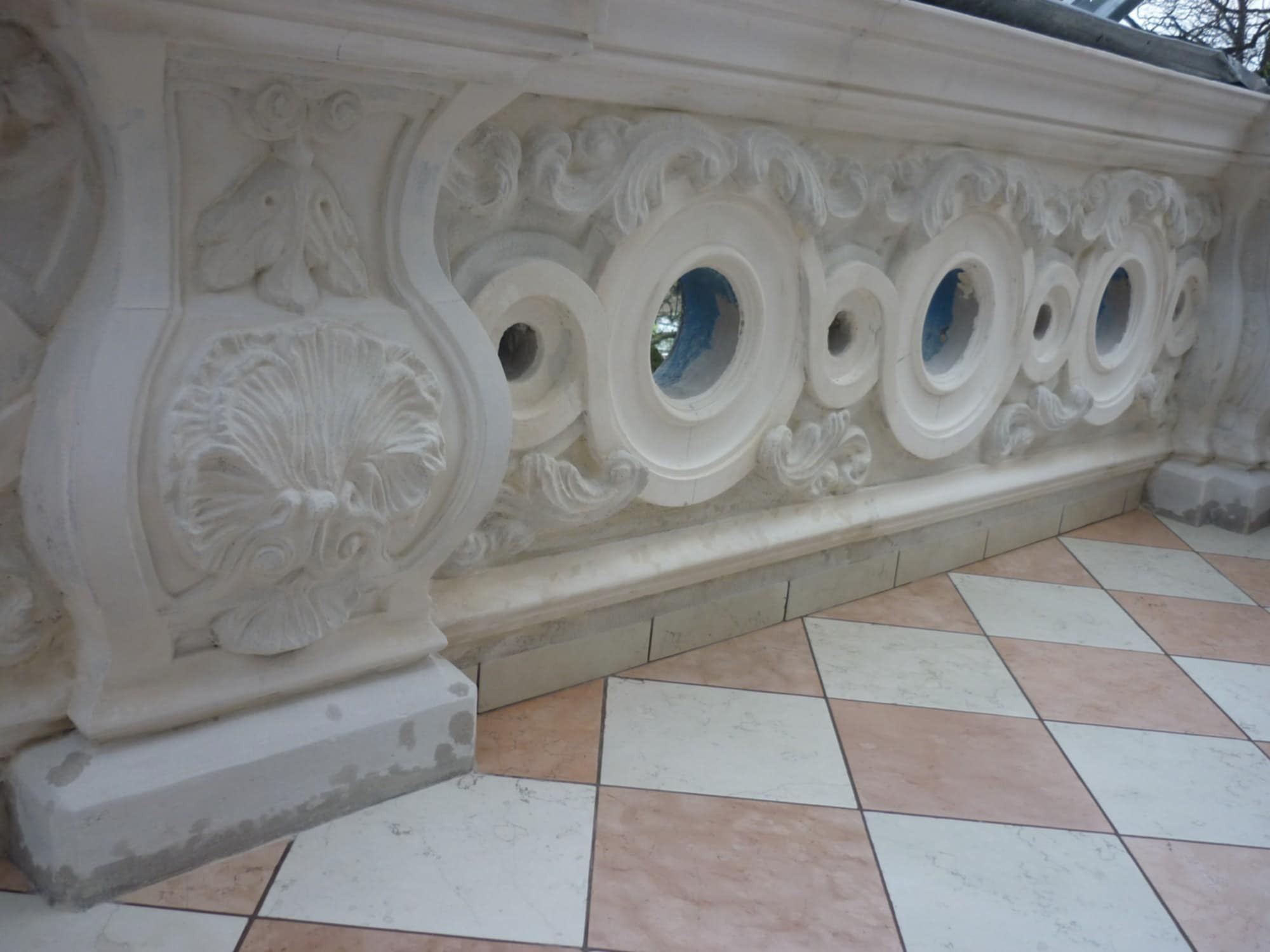 Biała, zdobiona balustrada z ornamentami i okrągłymi otworami na tle podłogi w szachownicę. Element dekoracyjny w stylu klasycznym lub zabytkowym.